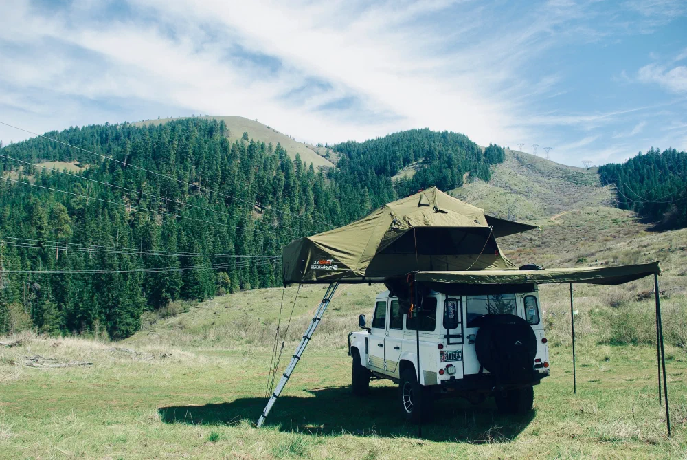 Rear three-quarter view of the Perentie 110 with rooftop tent and awning deployed in a mountain valley